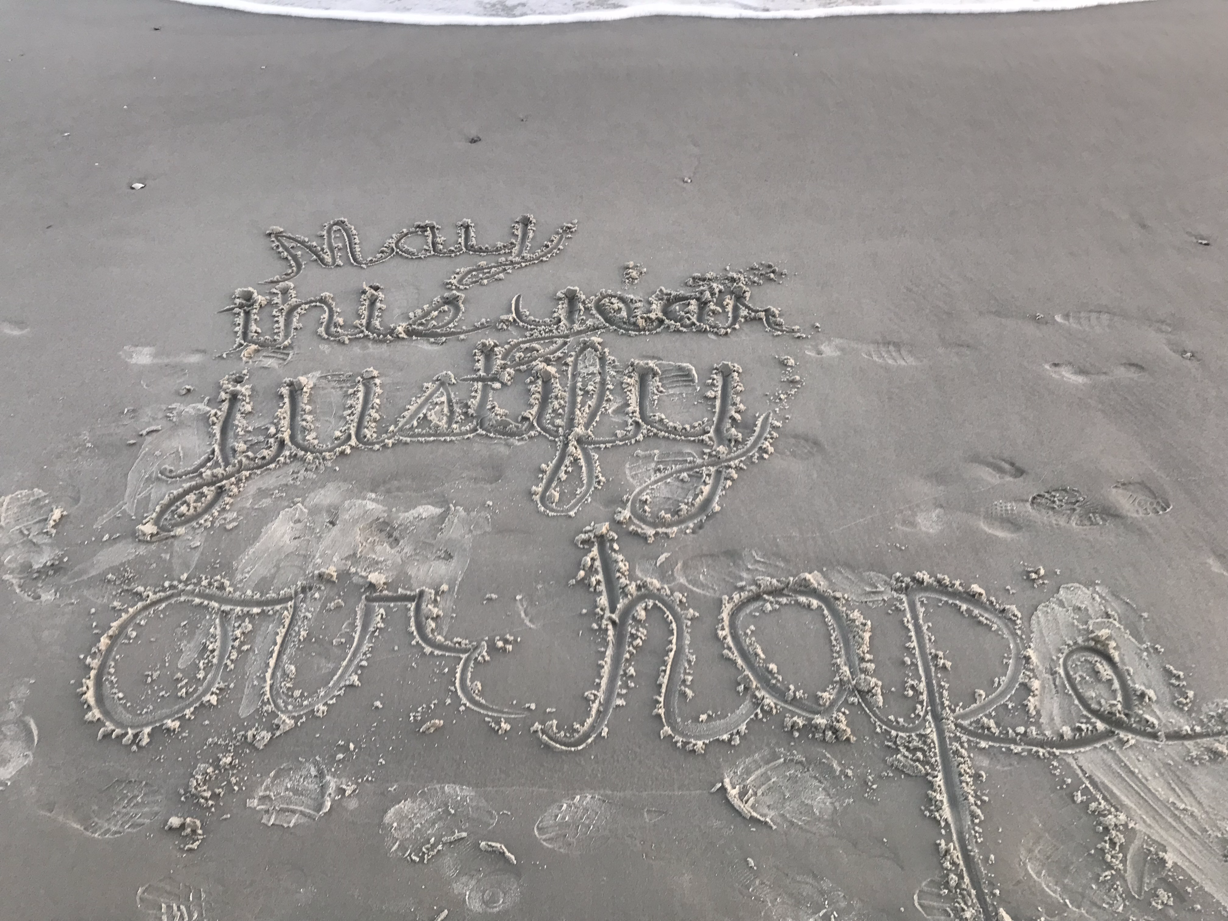 The phrase "May this year justify our hope" written in cursive in the sand on a beach